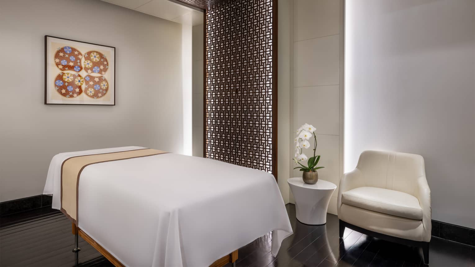 Massage table covered with white cloth in the centre of a treatment room with light-grey walls, a patterned wall accent, a painting and a white table and arm chair to the side