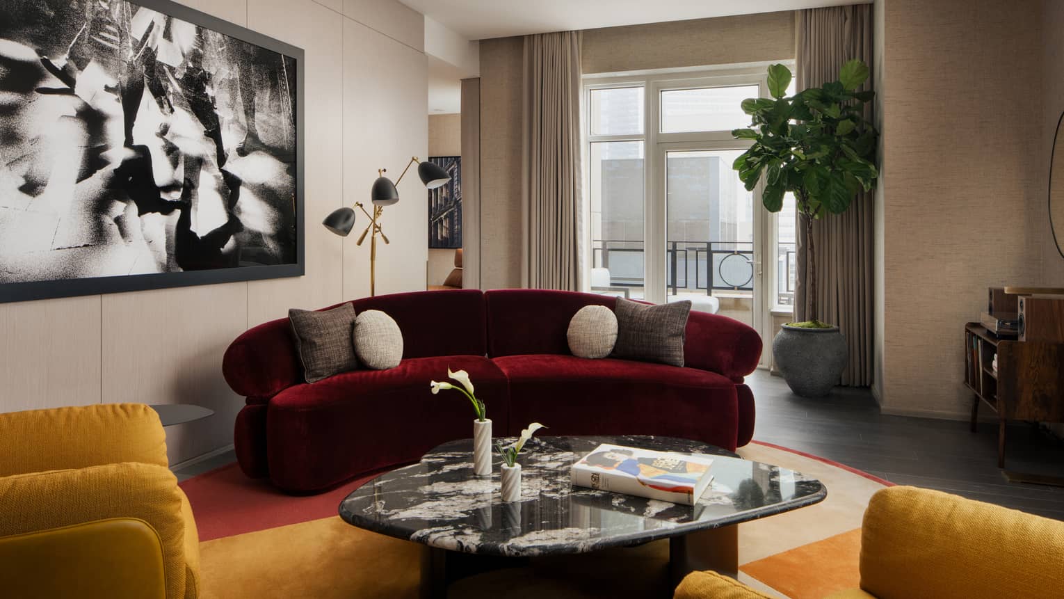 Luxury suite living room with burgundy sofa, coffee table, two yellow armchairs, large black-and-white wall art, potted plant and city-view balcony
