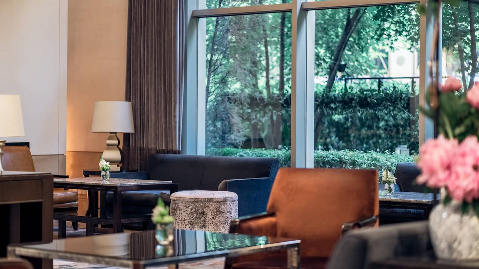 Glass-topped tables, blue couch, tan accent chair, floor-to-ceiling windows with open curtains displaying green foliage outside