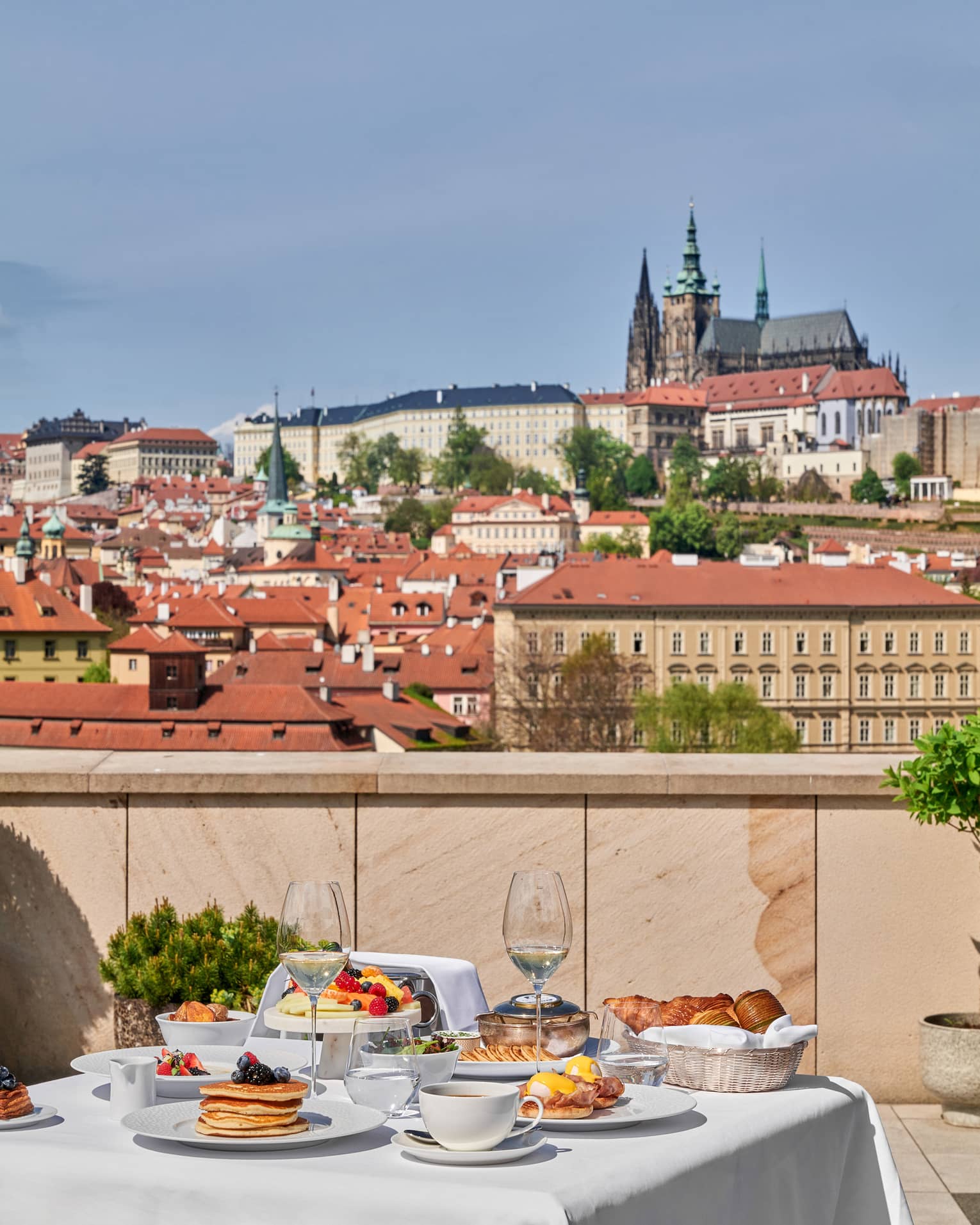 A white cloth?covered dining table on a terrace, set for a meal, with city views in the background