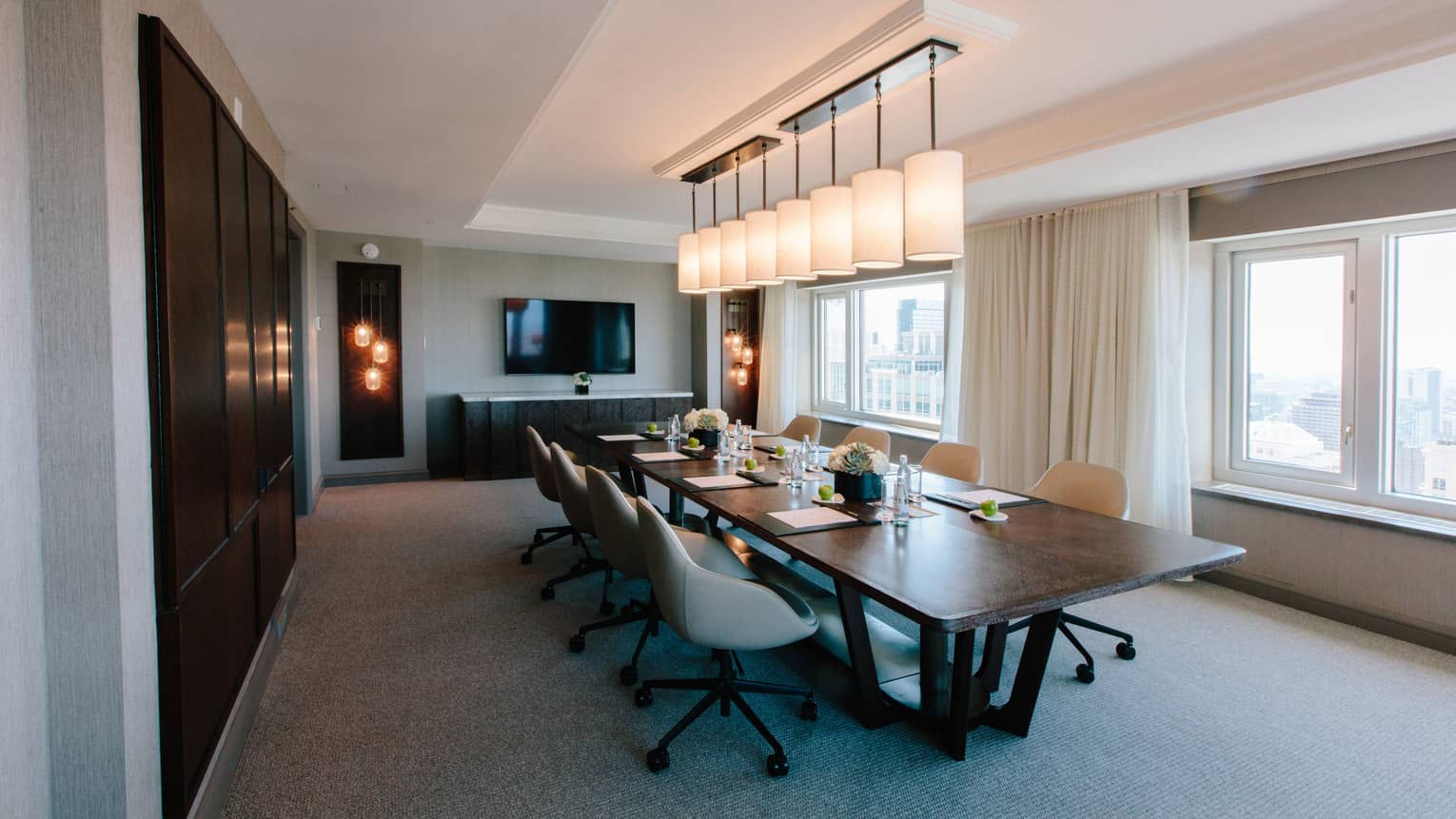 A meeting room with overhead lights, windows and a long wooden table.
