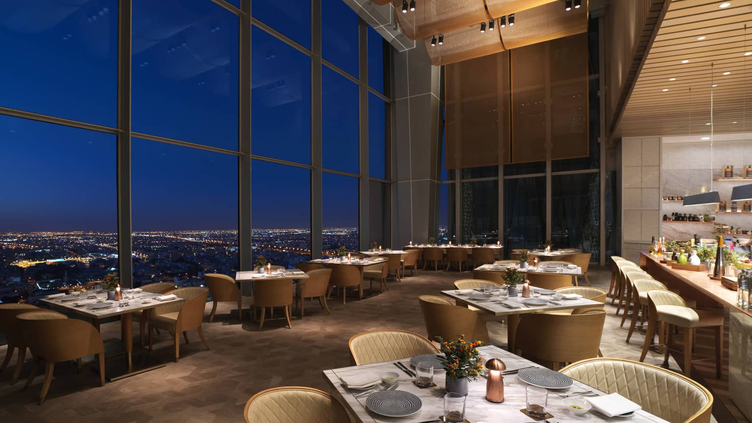 A dimly lit room with with a series of dining tables set up next to floor to ceiling windows.