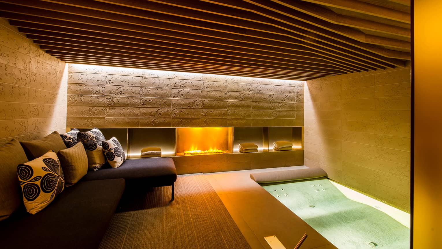 Gas fireplace in under stone wall in Urban Spa, small L-shaped sofa, cushions, spa tub