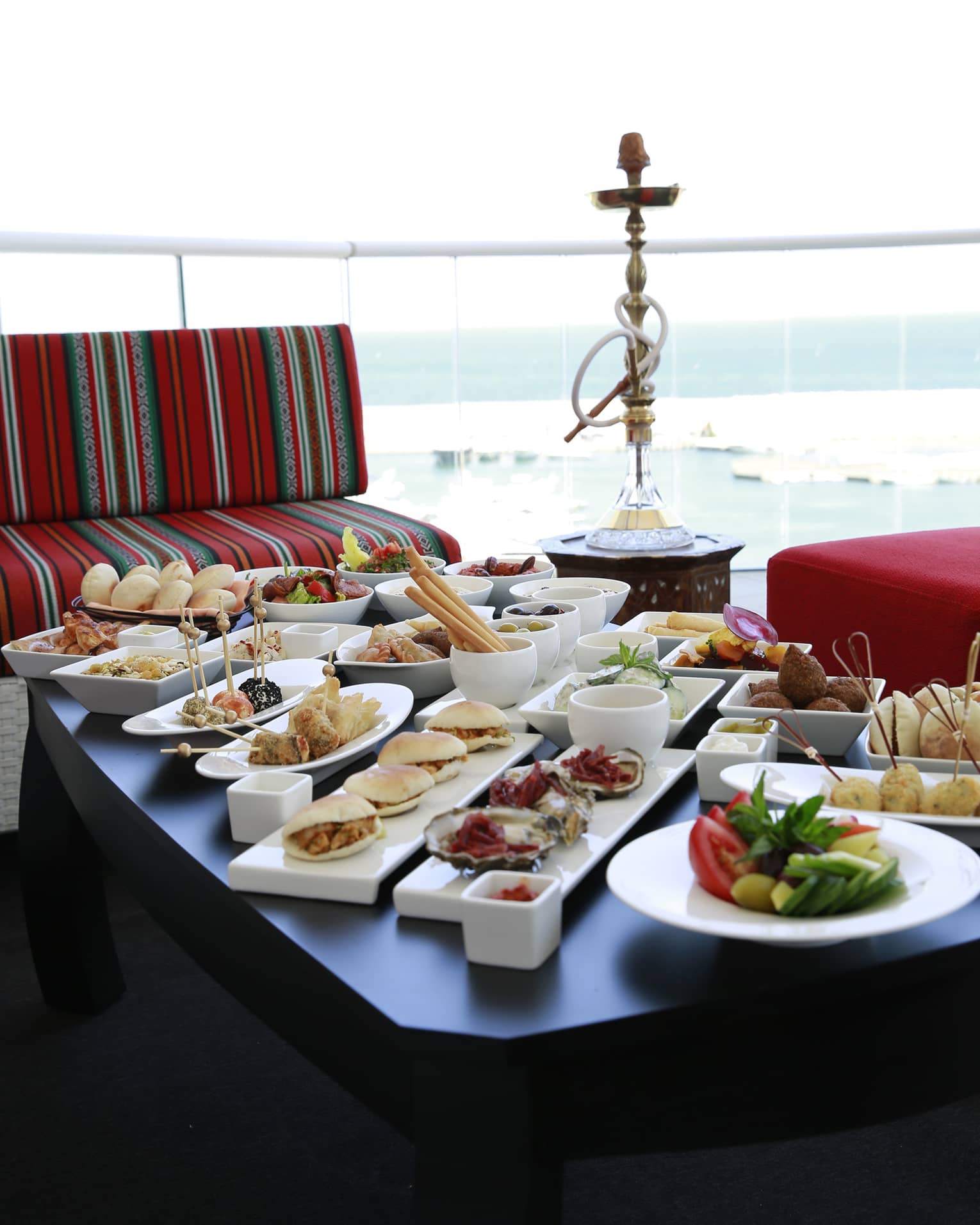 Table with more than a dozen white dishes with mezze appetizers and snacks, striped sofa and red bench