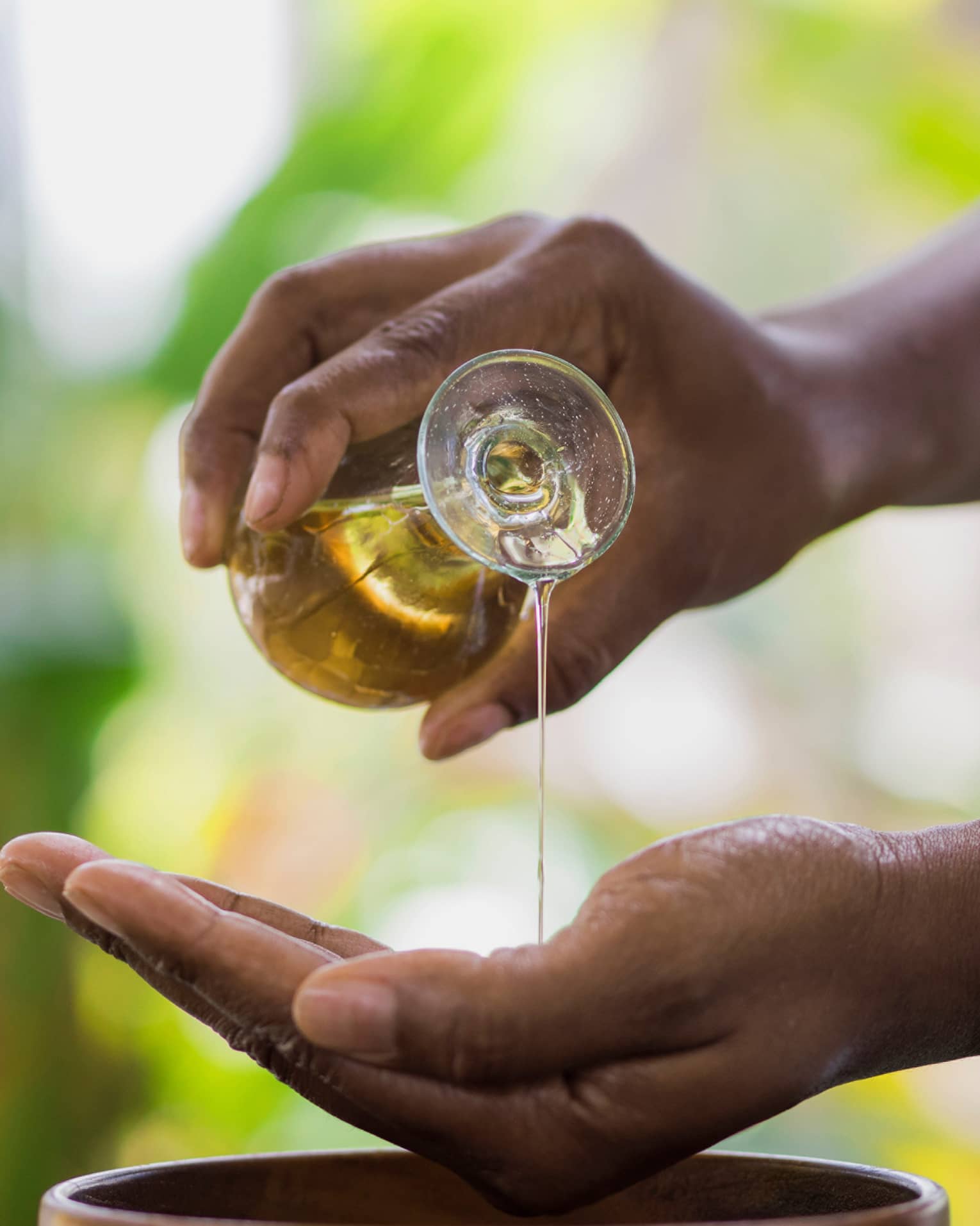 Golden oil flows from a small carafe held in one hand onto the other, a small wooden bowl underneath to catch the overflow.