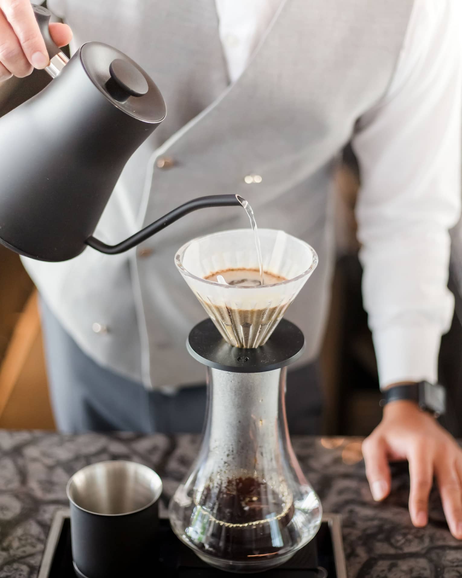 Barista holds black teapot, pours hot water over coffee in cone filter and glass jug on tray