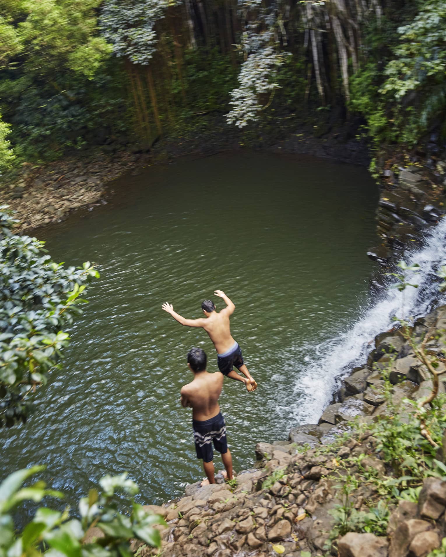 Two people leap from rocks to water below waterfall on the road to Hana