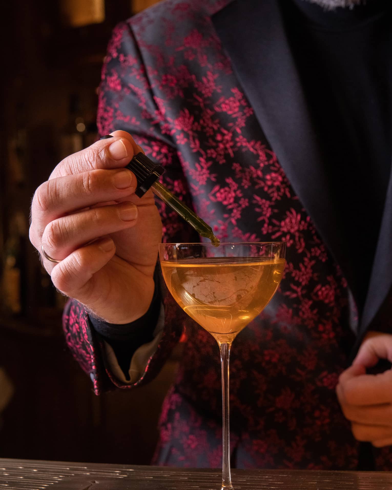 Bartender in red blazer uses dropper to add ingredient to bright-orange Basil cocktail in cordial glass