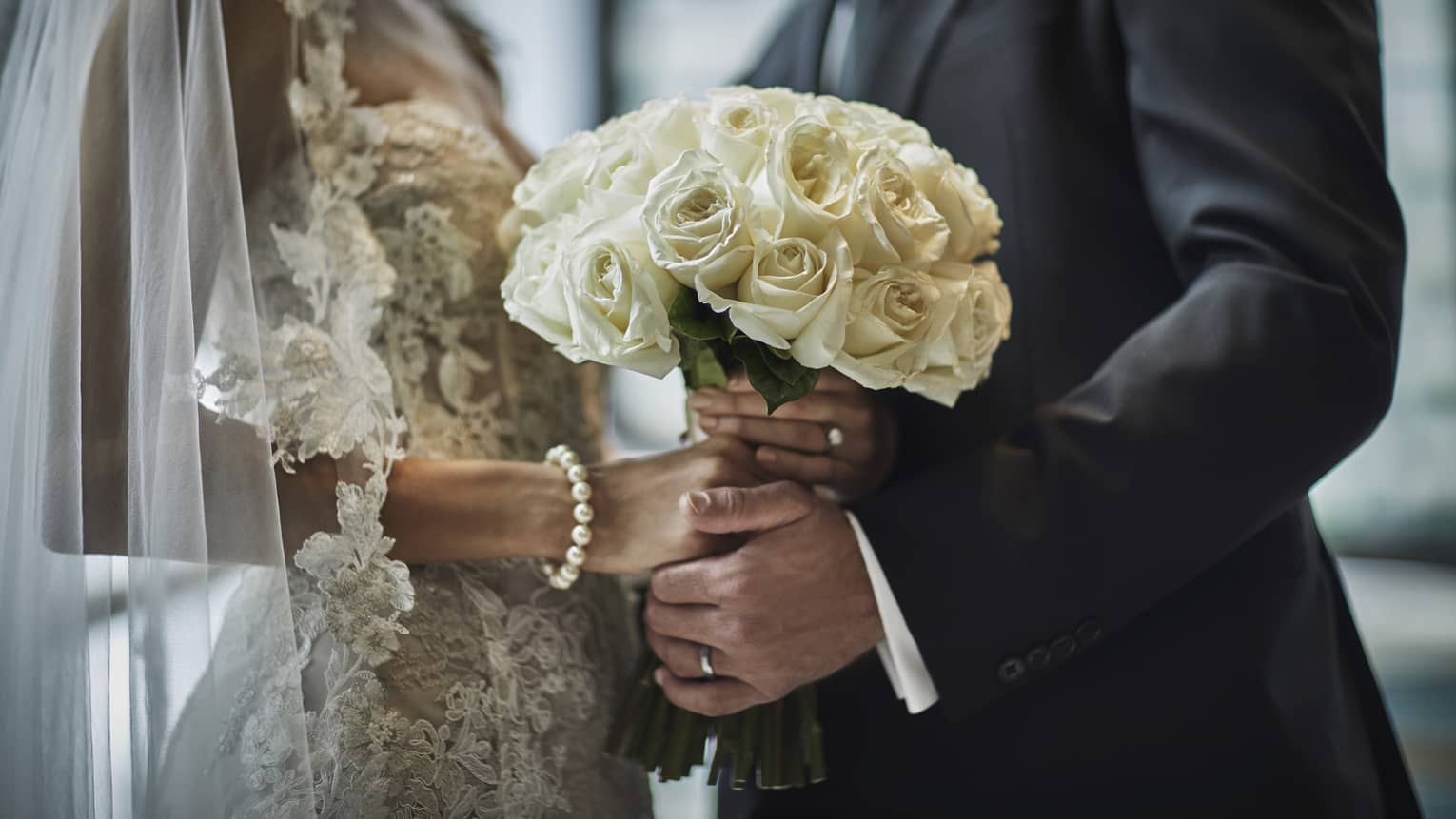 A close up of a bride in a lace gown and a groom in a gray suit hold the bride's bouquet of white roses together.
