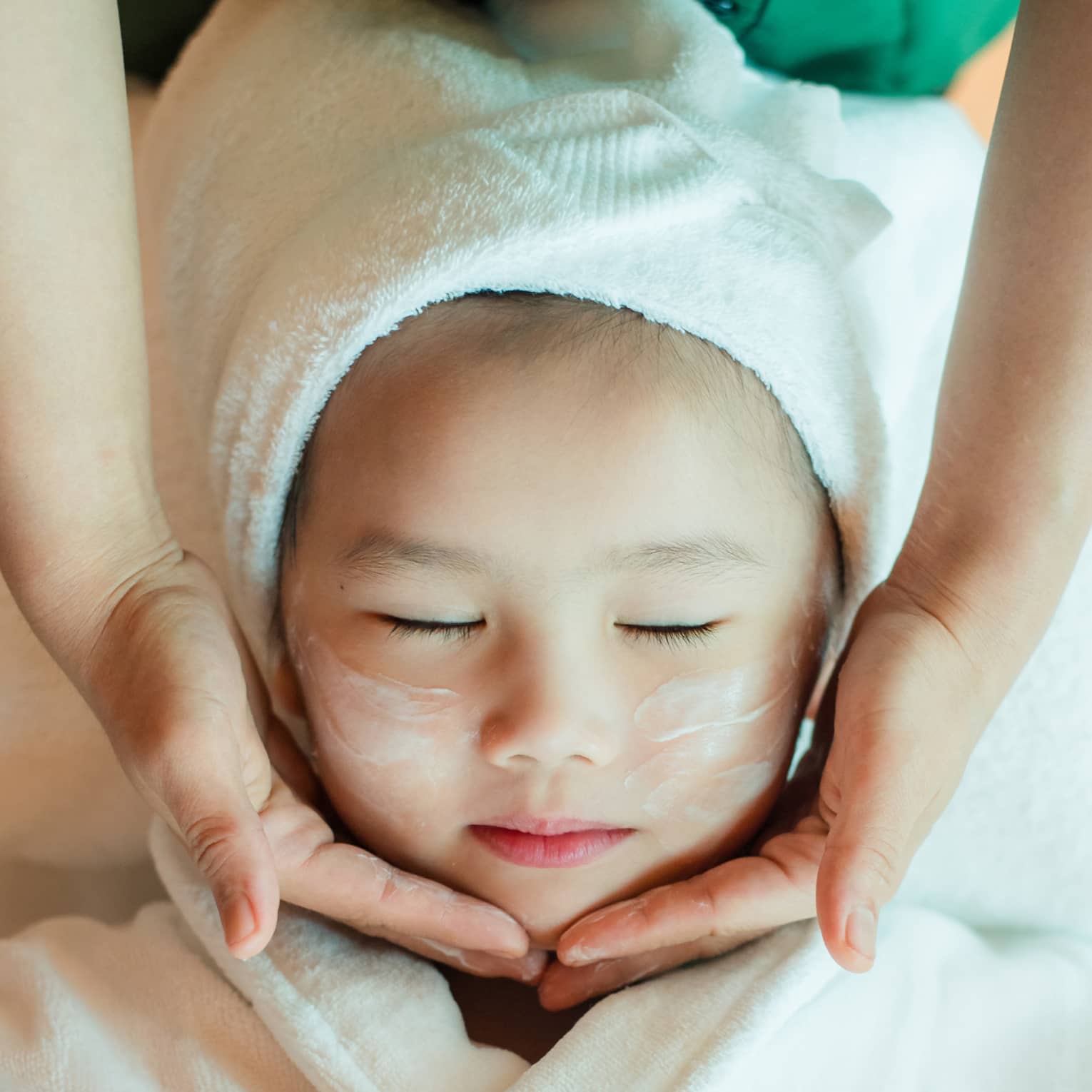 A close-up of a relaxed child wrapped in a towel and robe and enjoying a masseuse massaging a cream into their face.