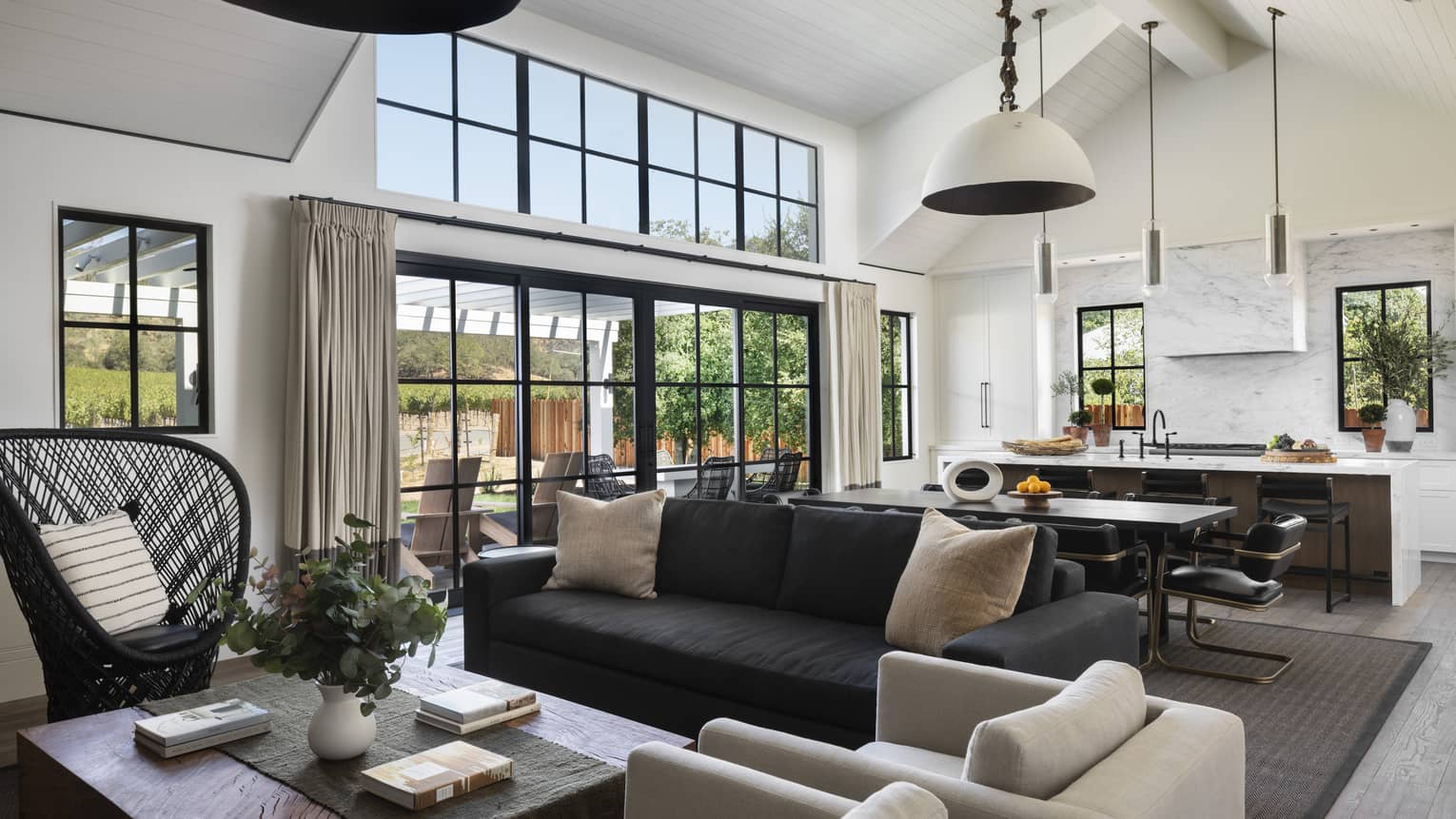 Open-concept living and kitchen area with a black sofa, beige armchairs, wicker chair, dining table, marble island and windows with views of the outdoor patio