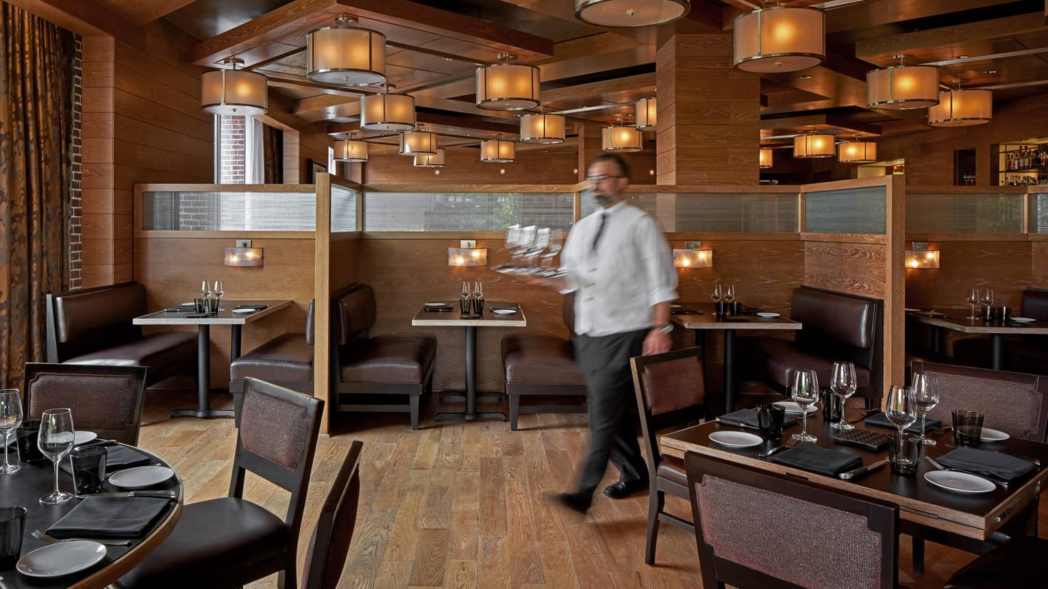 A team member walking with a tray of glasses through dimly lit restaurant with wooden floor and tables.