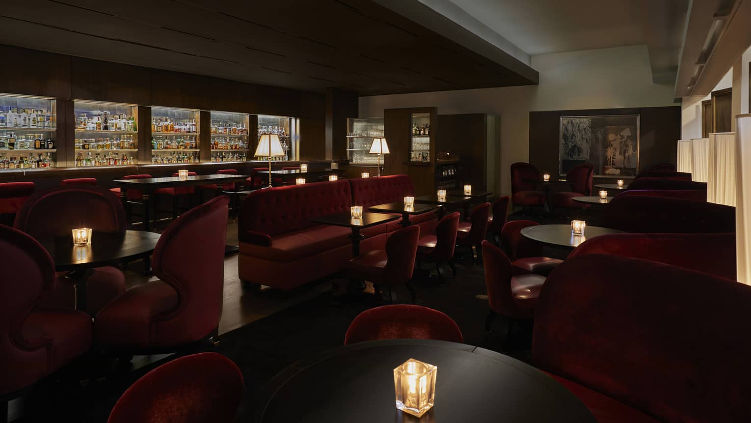 A dimly lit lounge with red chairs and a large selection of drinks behind a bar.