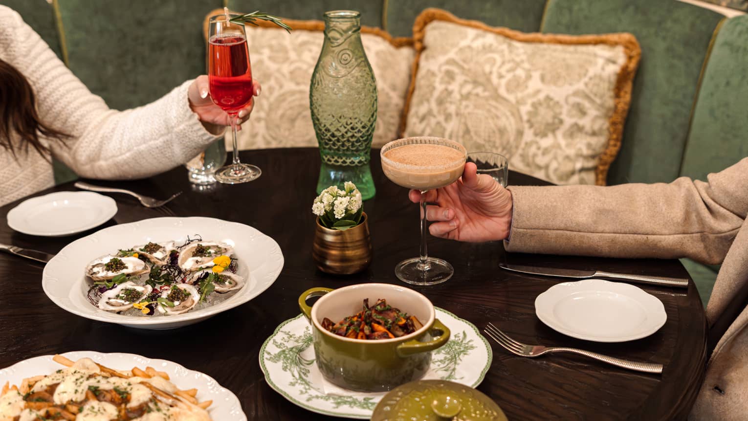 Two guests holding drinking glasses while seated at a dining table with multiple plates of food, including oysters.