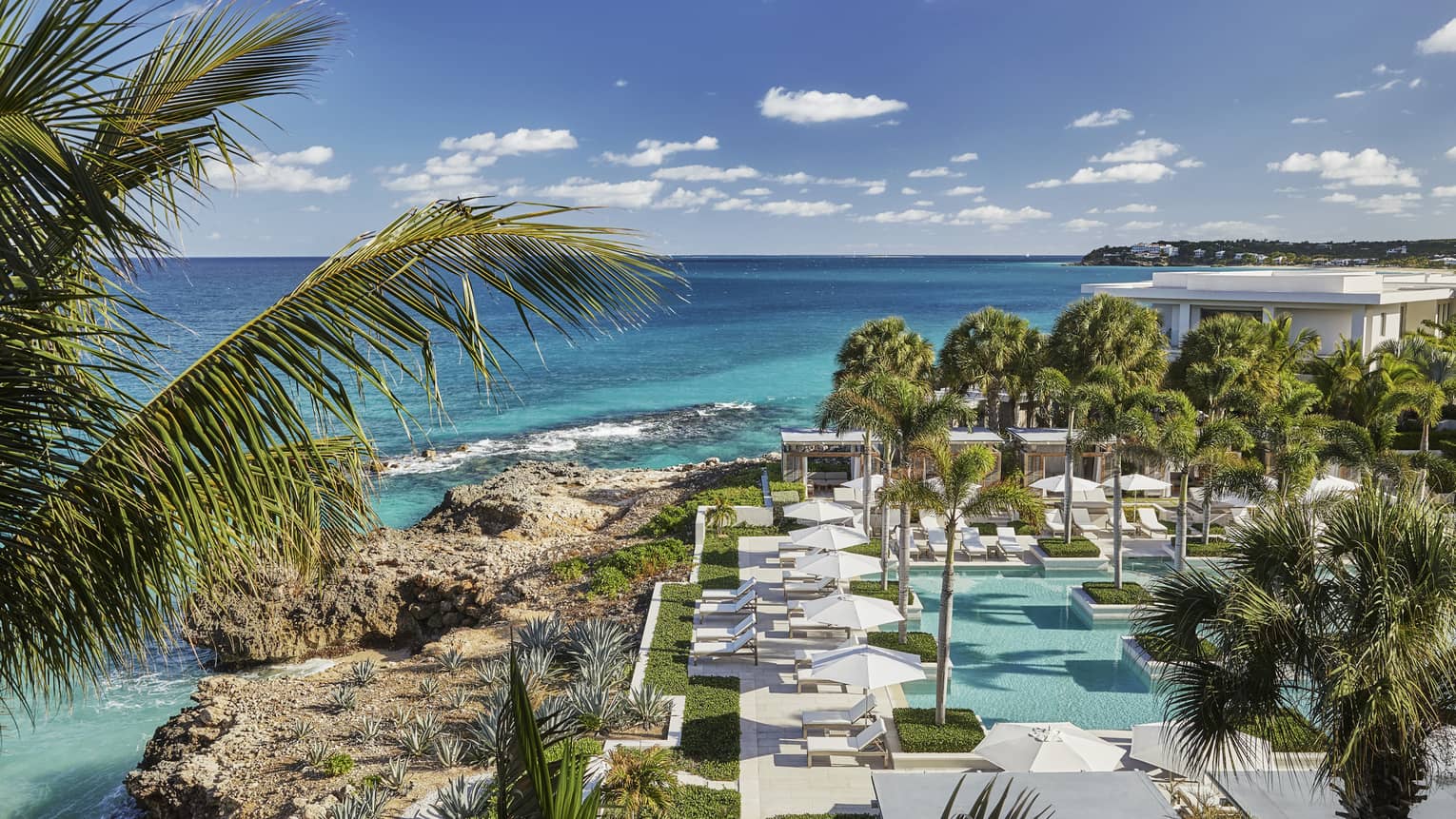 Looking down over Anguilla outdoor pool, patio, large rocks along blue-green ocean waterfront, blue sky