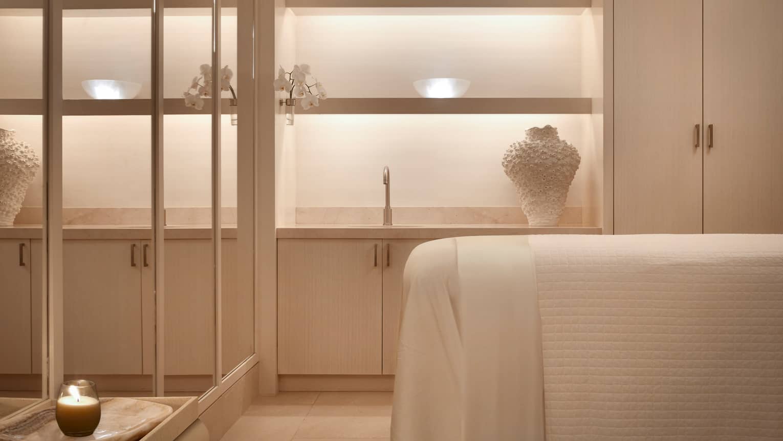 Spa treatment room bed with white linens across from mirror wall
