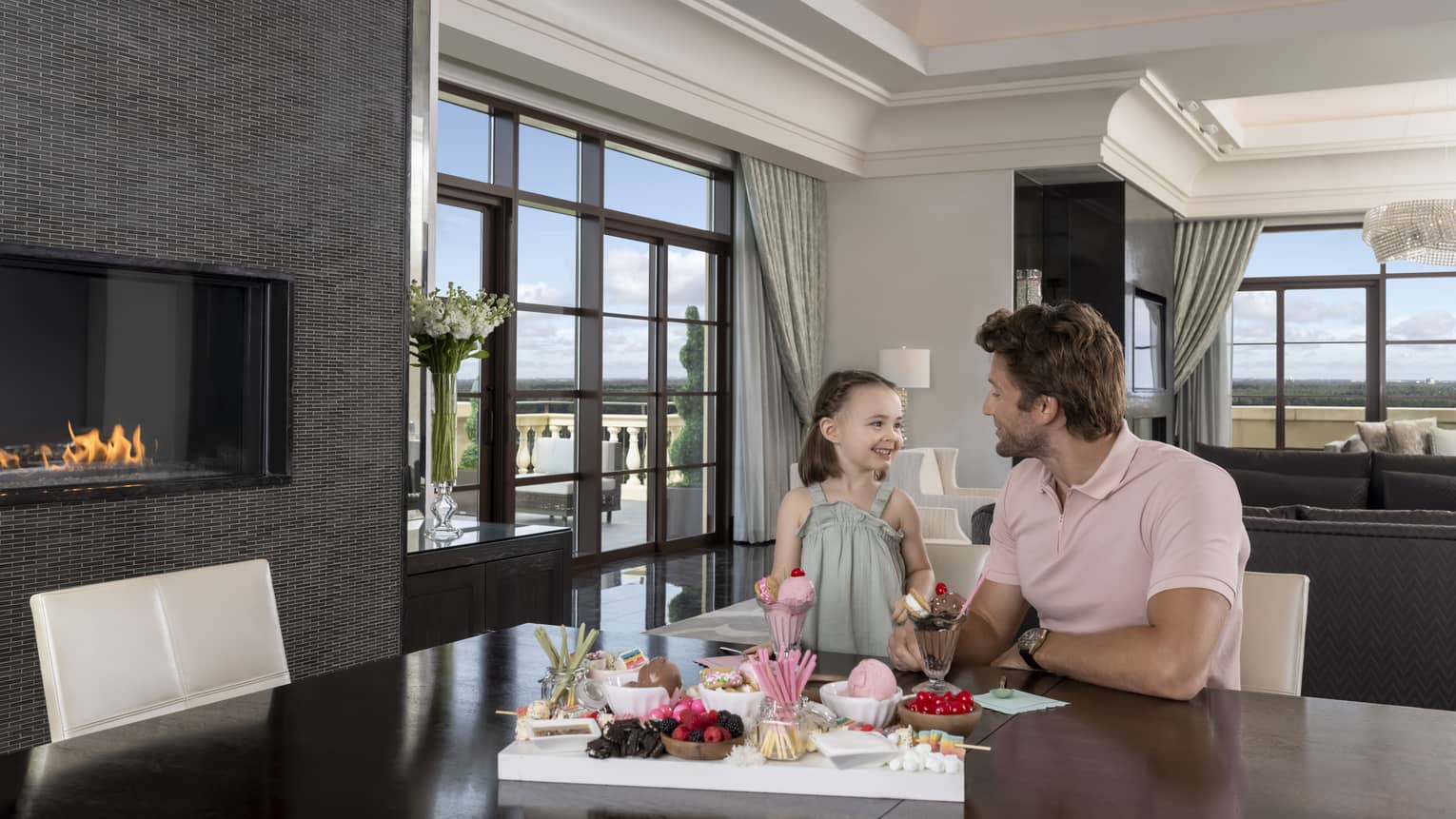 Man and young girl sit at dining table in hotel suite, next to fireplace
