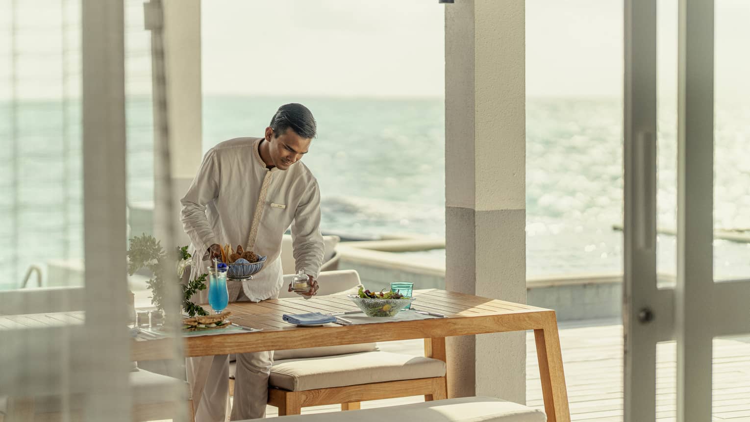 Male staff member sets table for In-Villa Dining, ocean in background