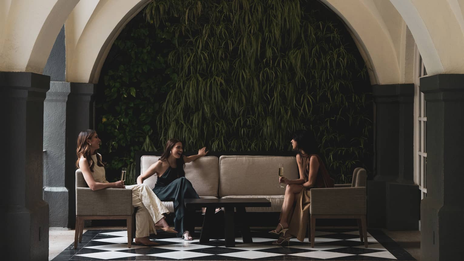 Three people sit on couches and chairs in a spacious seating area with an arched ceiling and a black-and-white tiled floor.