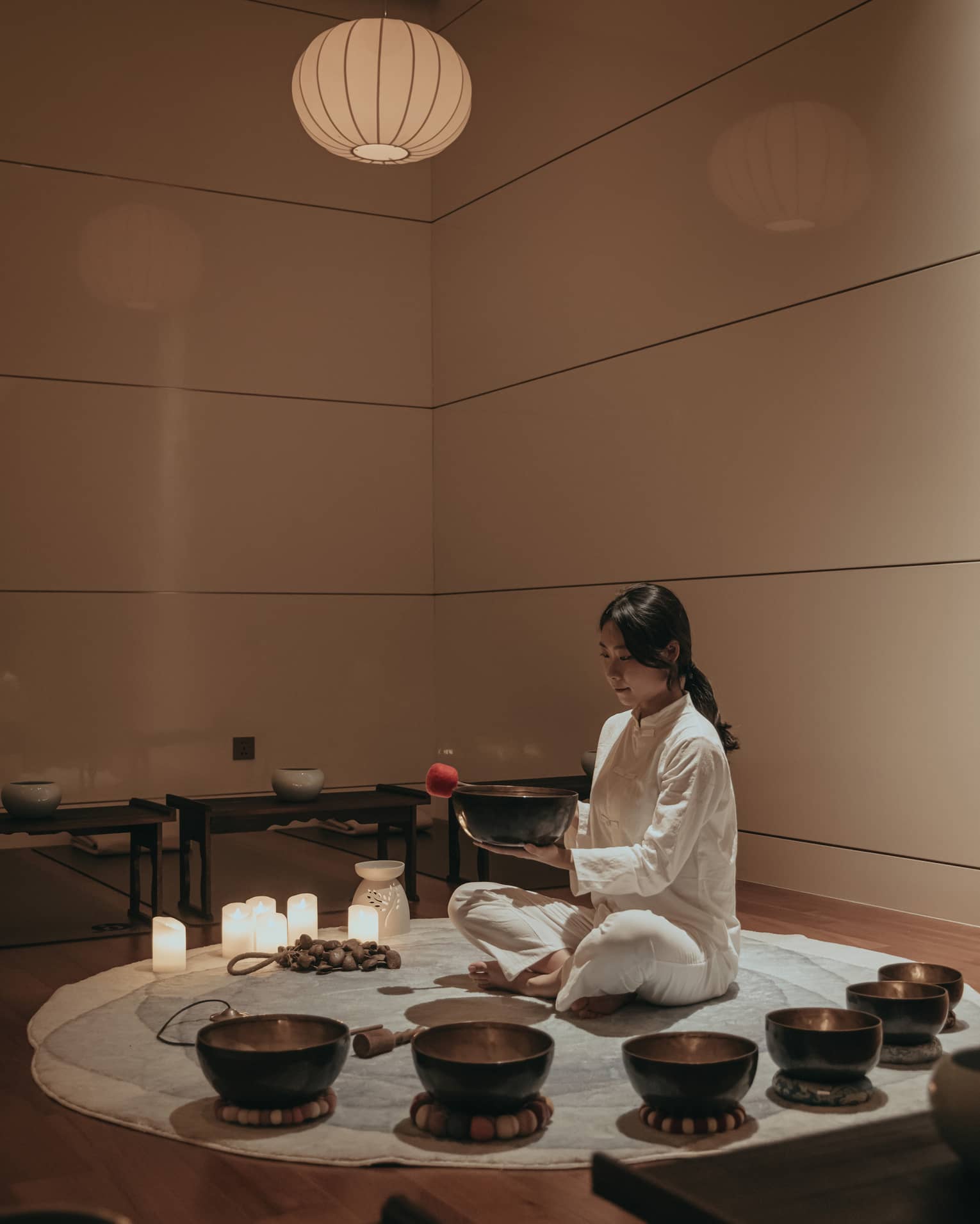 In a dim room, cross-legged on a circular rug amid various candles and bowls, a barefoot practitioner holds a bowl and mallet.
