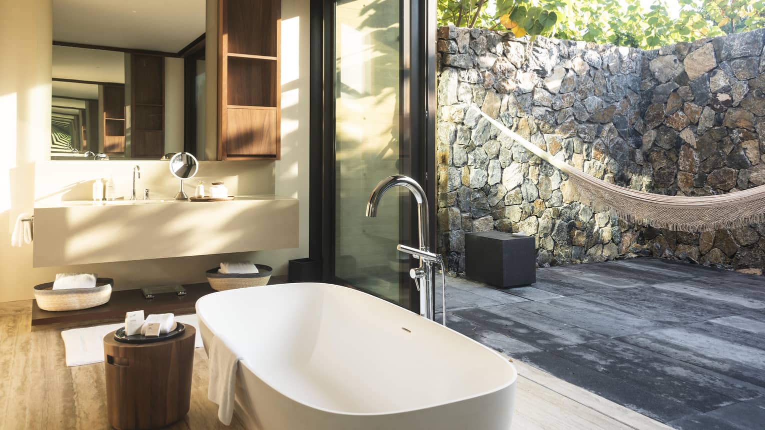 Modern bathroom with a free-standing tub, large vanity and floor-to-ceiling glass doors opening to a private outdoor patio with stone walls and a hammock