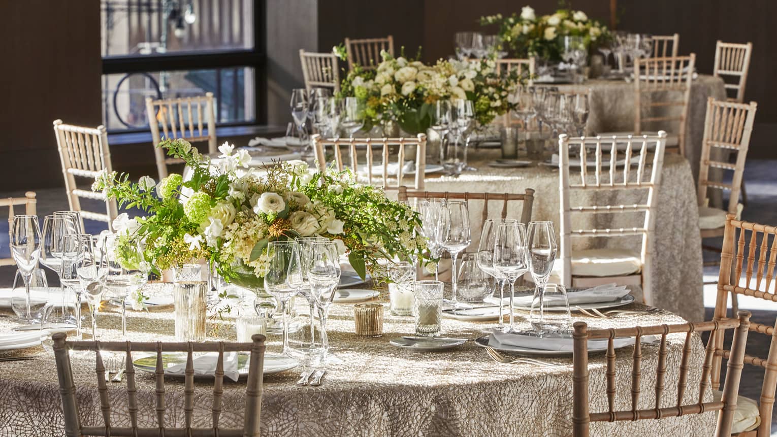 Elegantly set round wedding tables featuring white and gold linens, each enhanced by a display of fresh flower centerpieces and wooden chairs