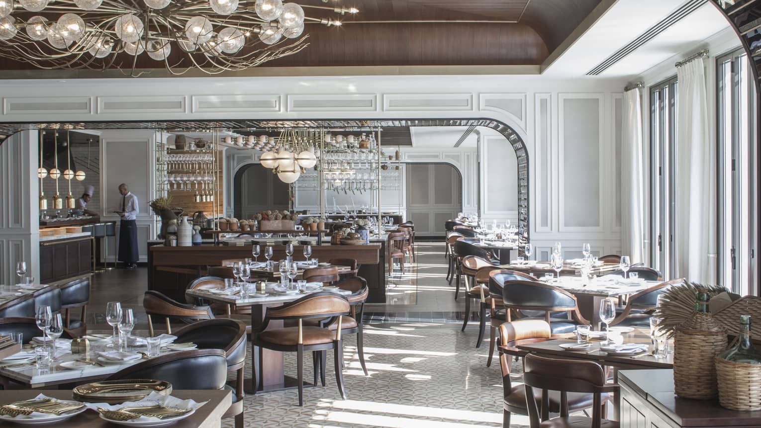 Large, sunny La Capitale dining room with elegant brown leather chairs, tables, white tile, modern chandeliers, server at counter