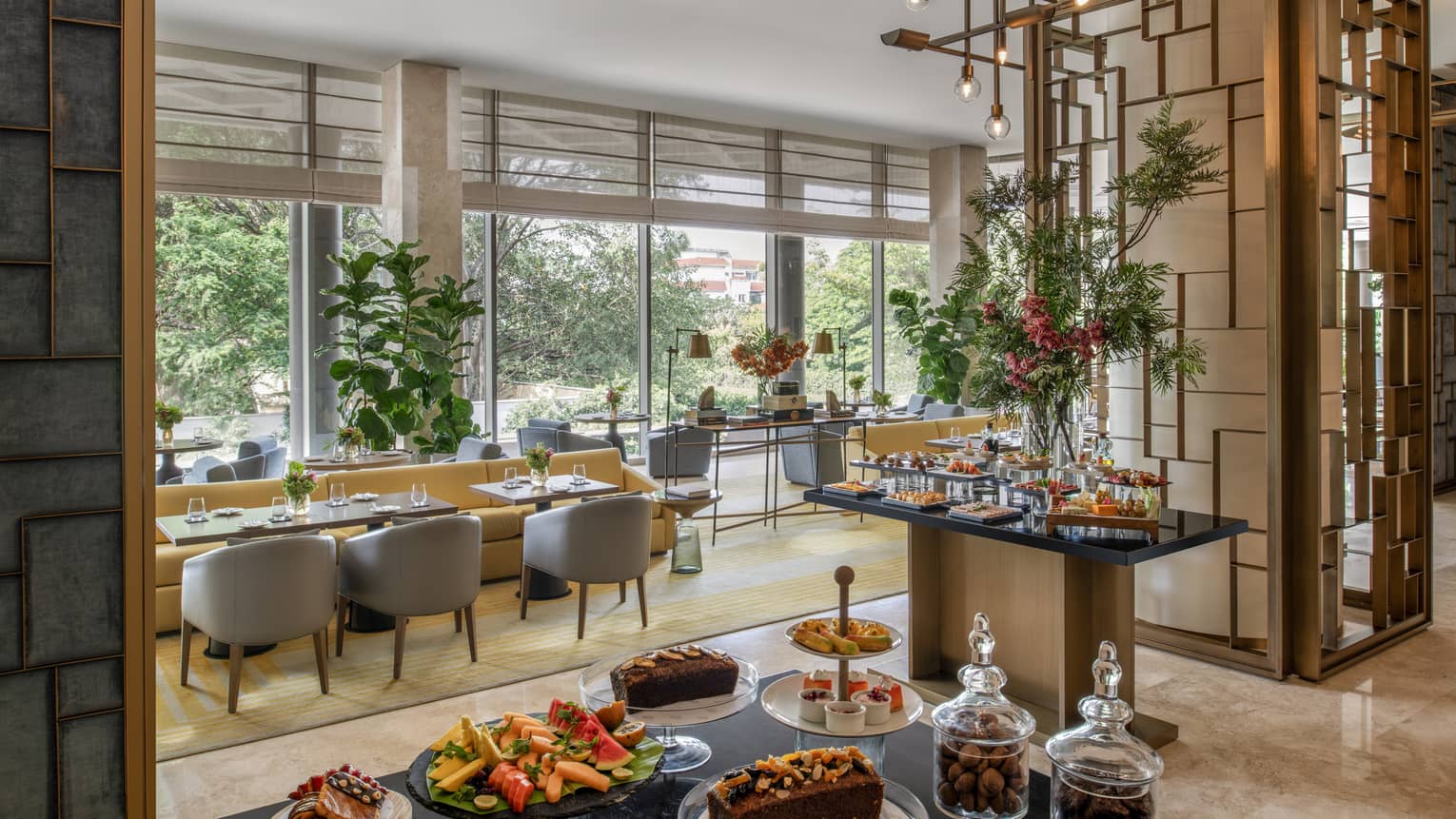 Brightly lit room with displays of food, long rectangular tables and grey arm chairs, ornate metal wall art