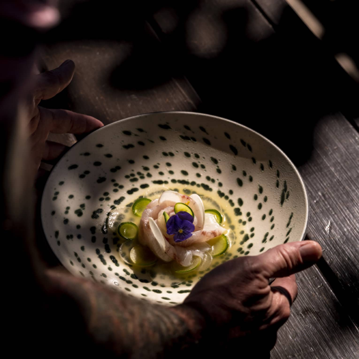 An artistically arranged seafood dish topped with a pansy flower is gently placed on a wooden table dappled with light.