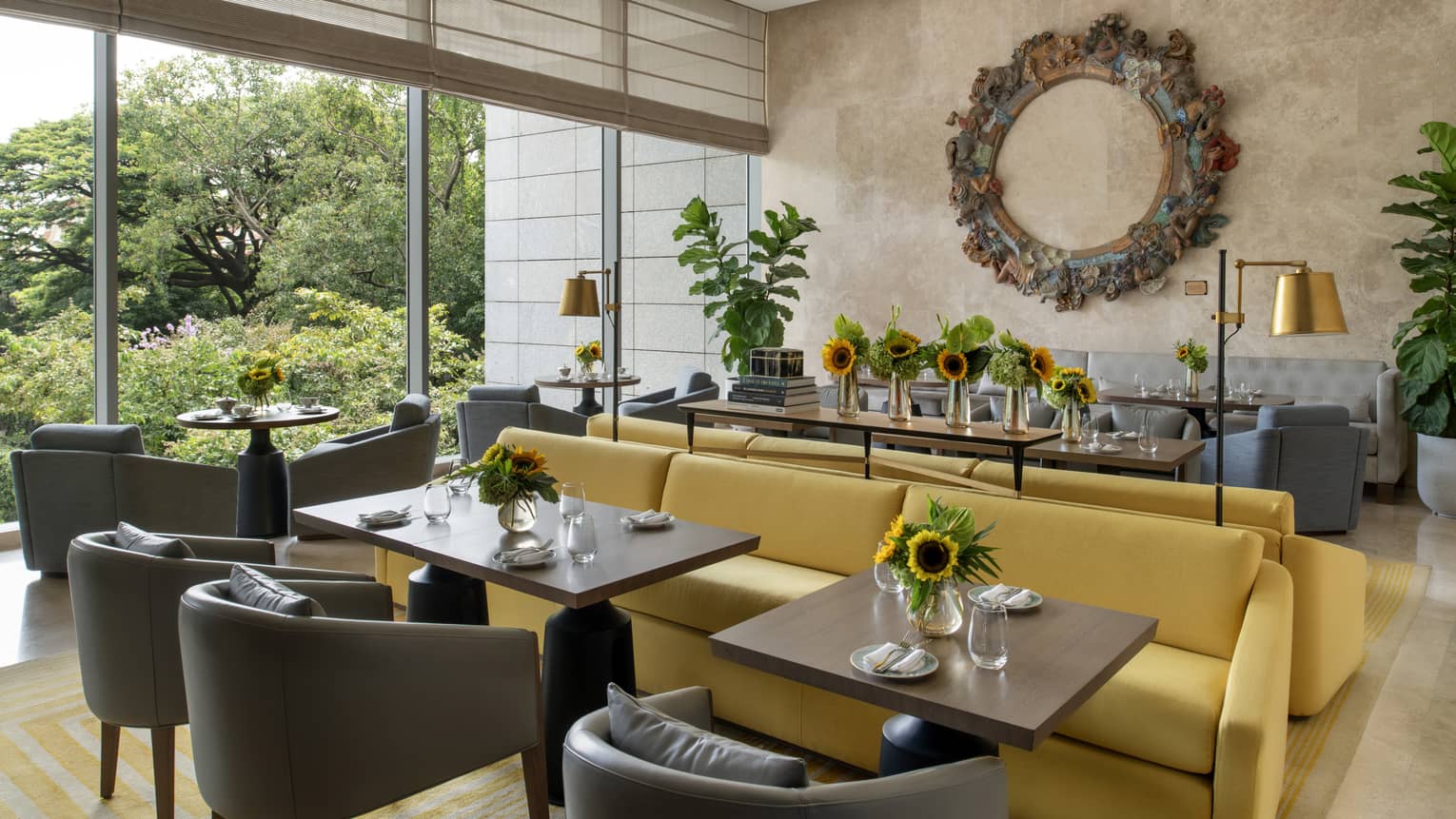 Brightly lit restaurant with yellow couches and grey armchairs, sunflower centrepieces and ornate metal wall piece