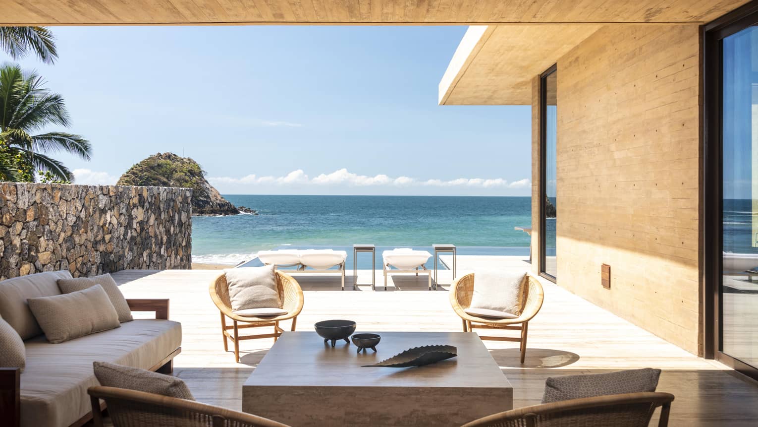 Outdoor seating area with wooden furniture and wicker chairs overlooking a beachfront view. Lounge chairs are positioned by the water with a rocky island in the distance.