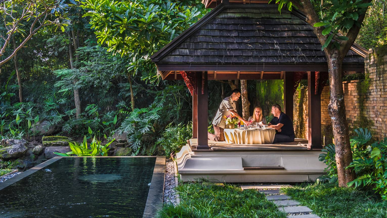 Hotel staff pours fresh juice for couple at private patio dining table under wood gazebo