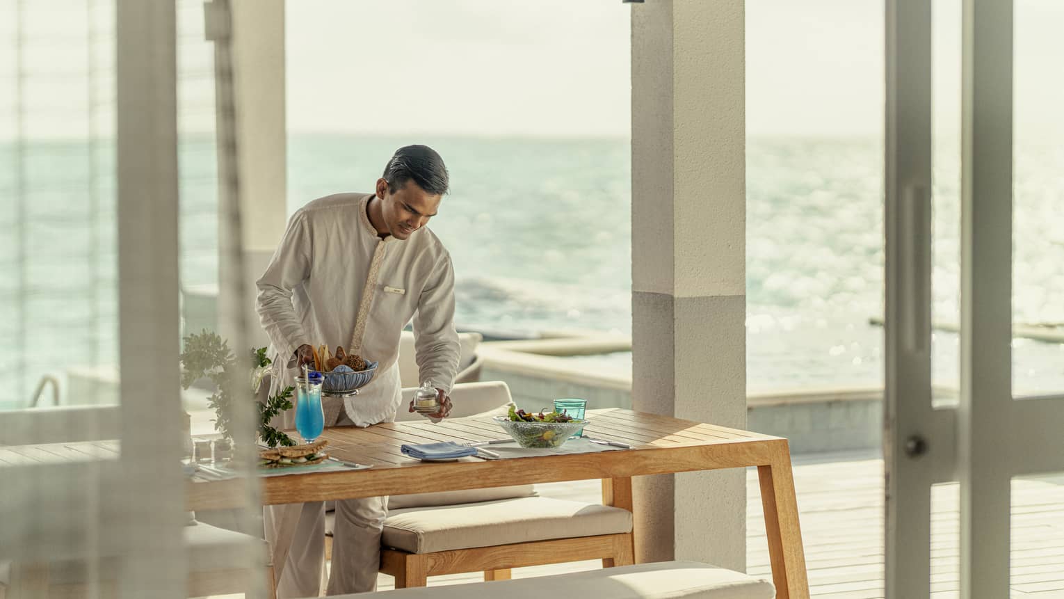 Male staff member sets table for In-Villa Dining, ocean in background