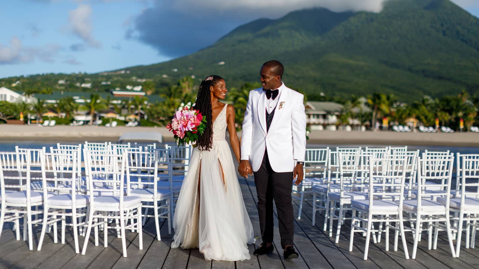 Woman in wedding dress, pink bouquet, holding hand of man in white jacket, black pants, chairs surrounding
