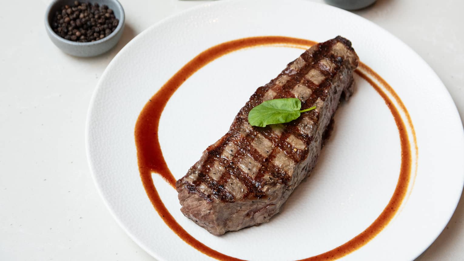 Strip steak topped with a green leaf for garnish and plated on a white round plate with a streak of sauce surrounding the steak