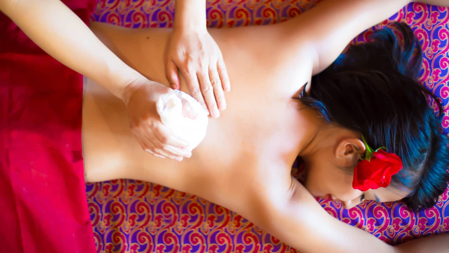 Masseuse rubs woman's bare back with sponge as she lies face-down on massage table with colourful cloths