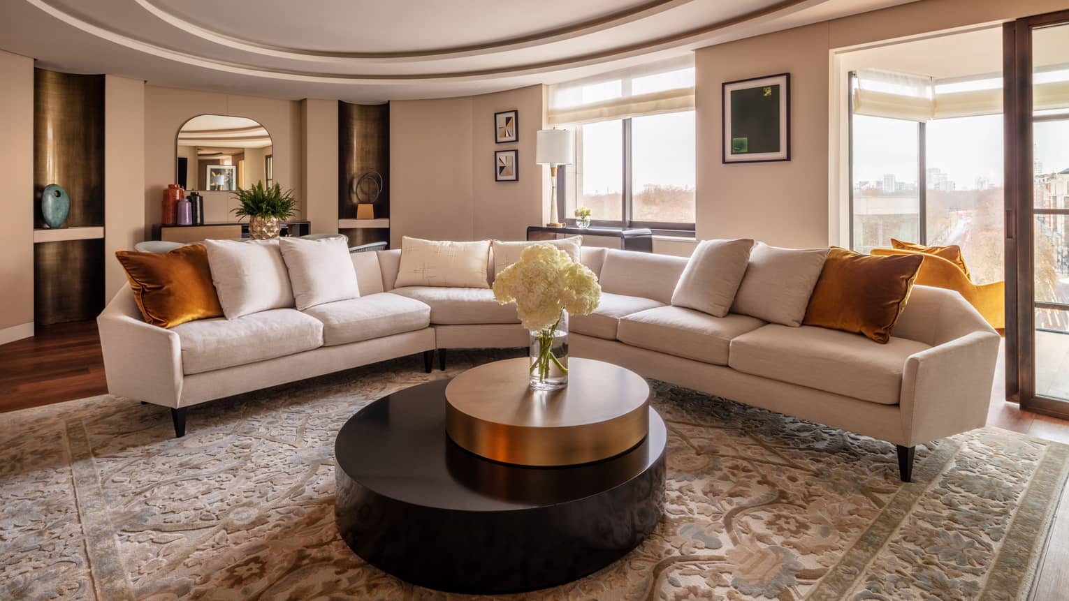 The Hyde Park Suite at Four Seasons London Park Lane in Mayfair showcases a sophisticated living room with a panoramic view of the city. The room features a long, sleek white sofas with orange throw pillows, arranged around a large circular black coffee table with a gold rim. The area is adorned with a luxurious beige patterned rug, adding texture and warmth. Artworks hang on the walls, and a large window frames the stunning view outside, providing ample natural light. The decor is elegantly completed with a neutral colour palette and modern furnishings, creating an inviting and stylish space.