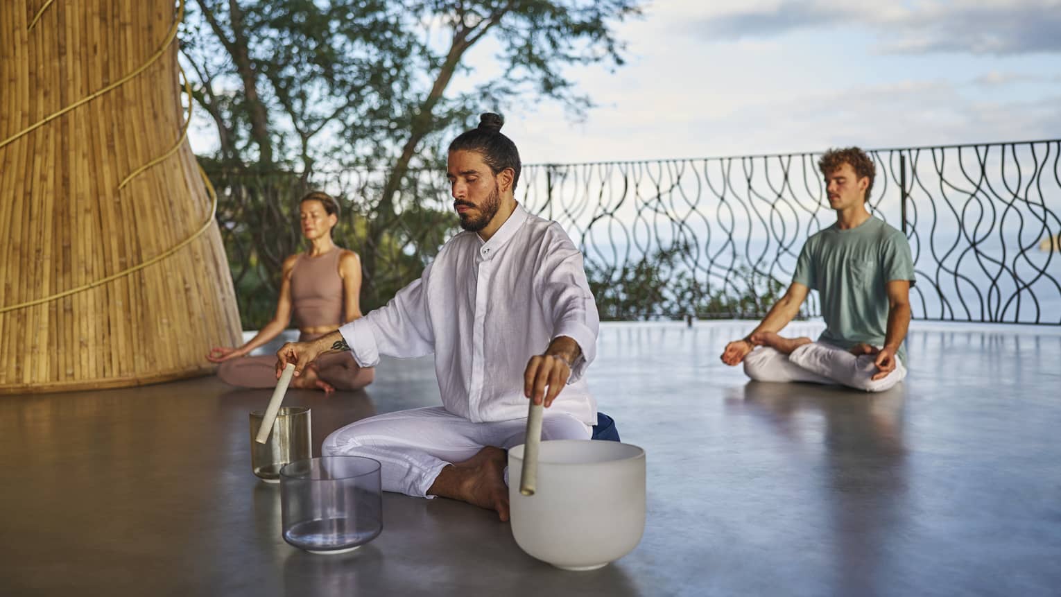 Man wearing all white clothing sits cross-legged on floor with three singing bowls in front of him and two people also sitting cross-legged on the floor behind him