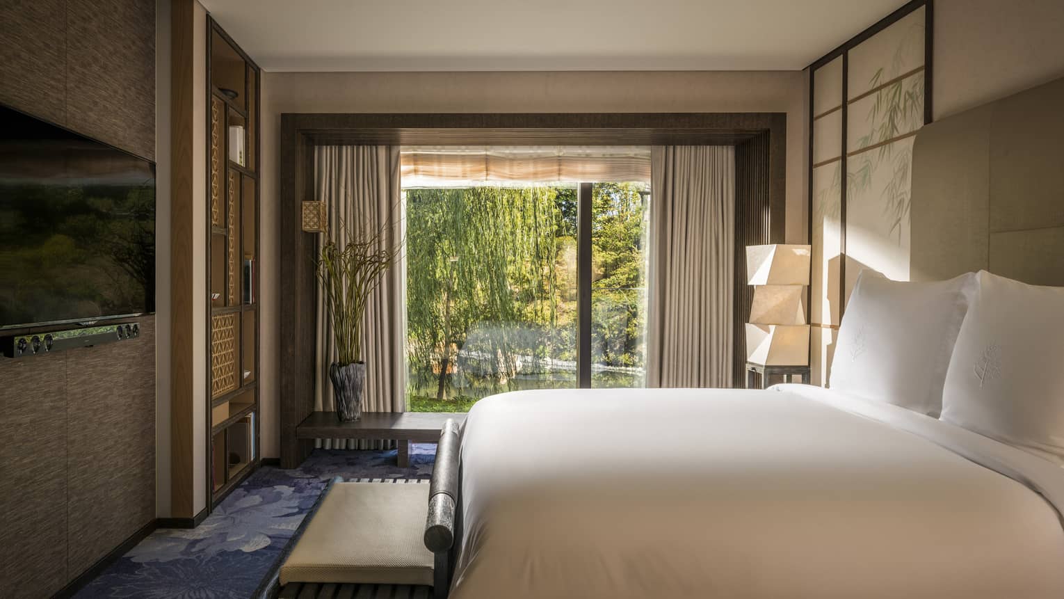 Hotel bedroom with a king bed facing TV, soft lighting and a large window offering a serene garden view