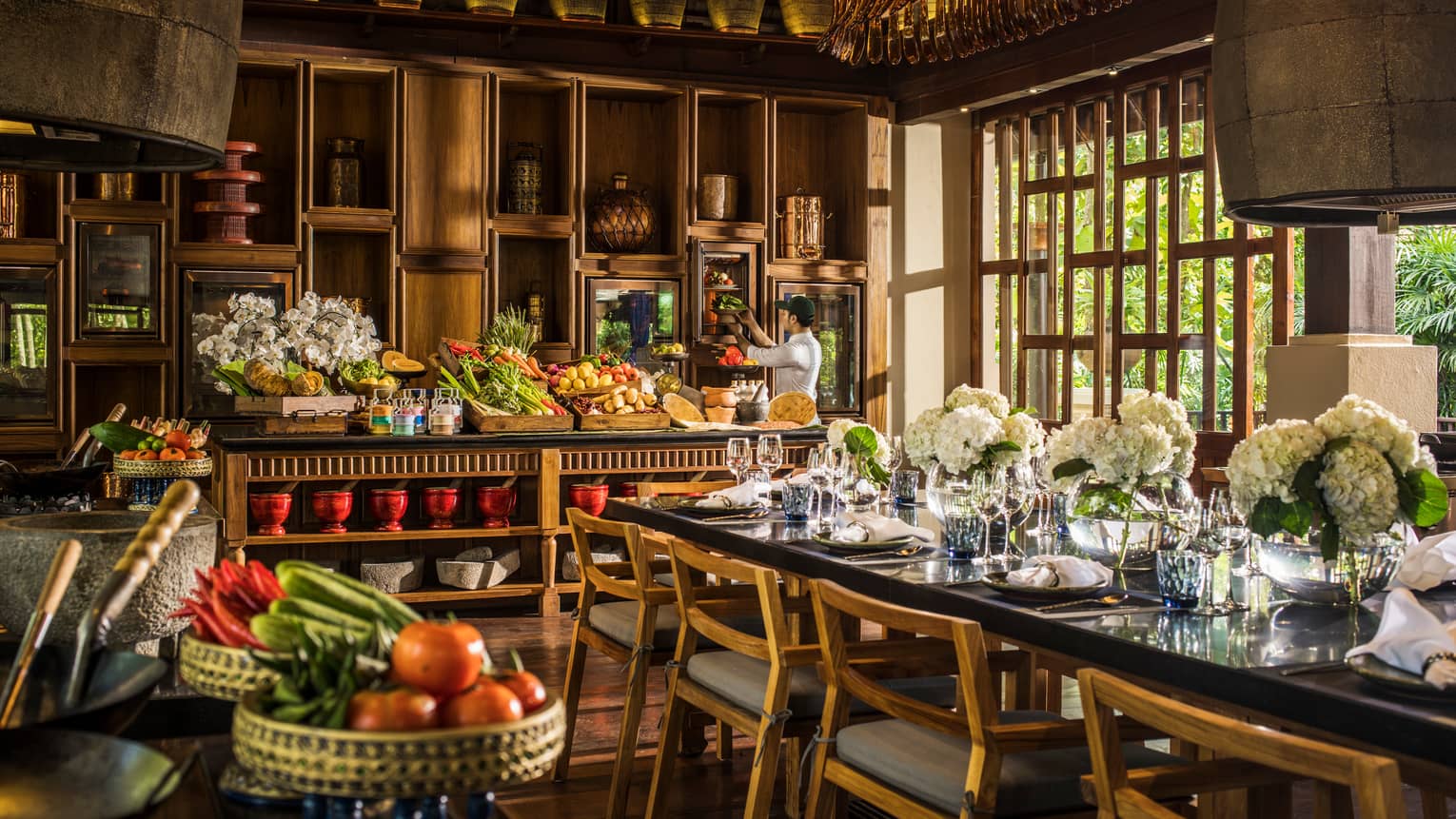Close-up of end of dining table with white flowers, kitchen line with baskets of fruits and vegetables