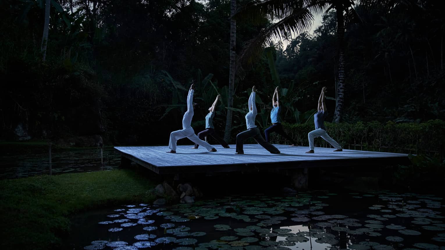 Five people kneel in yoga pose with arms above head on wood platform in forest at night