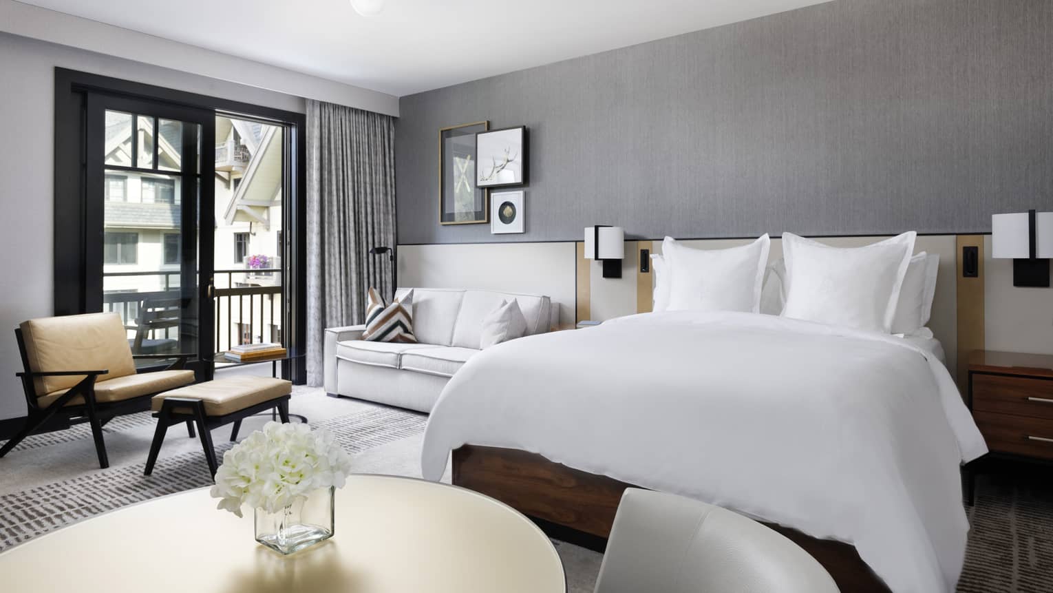 Guest room with King bed, white linens, small round dining table, white leather loveseat, arm chair, glass doors leading to balcony