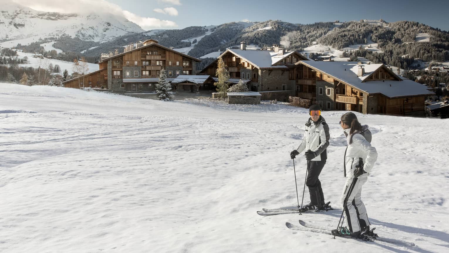 Man, woman on skis at top of snowy hill, Four Seasons Hotel Megeve in background