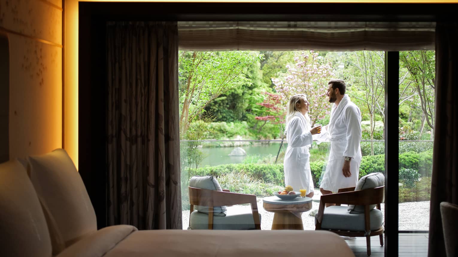 Smiling couple wearing white bathrobes on sunny balcony past hotel room bed
