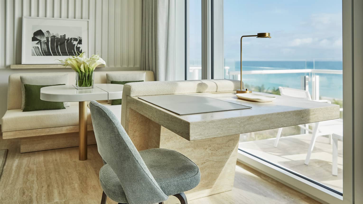 Luxury hotel room with a marble desk, a chair, a seating area with a table and flowers and floor-to-ceiling windows offering ocean views
