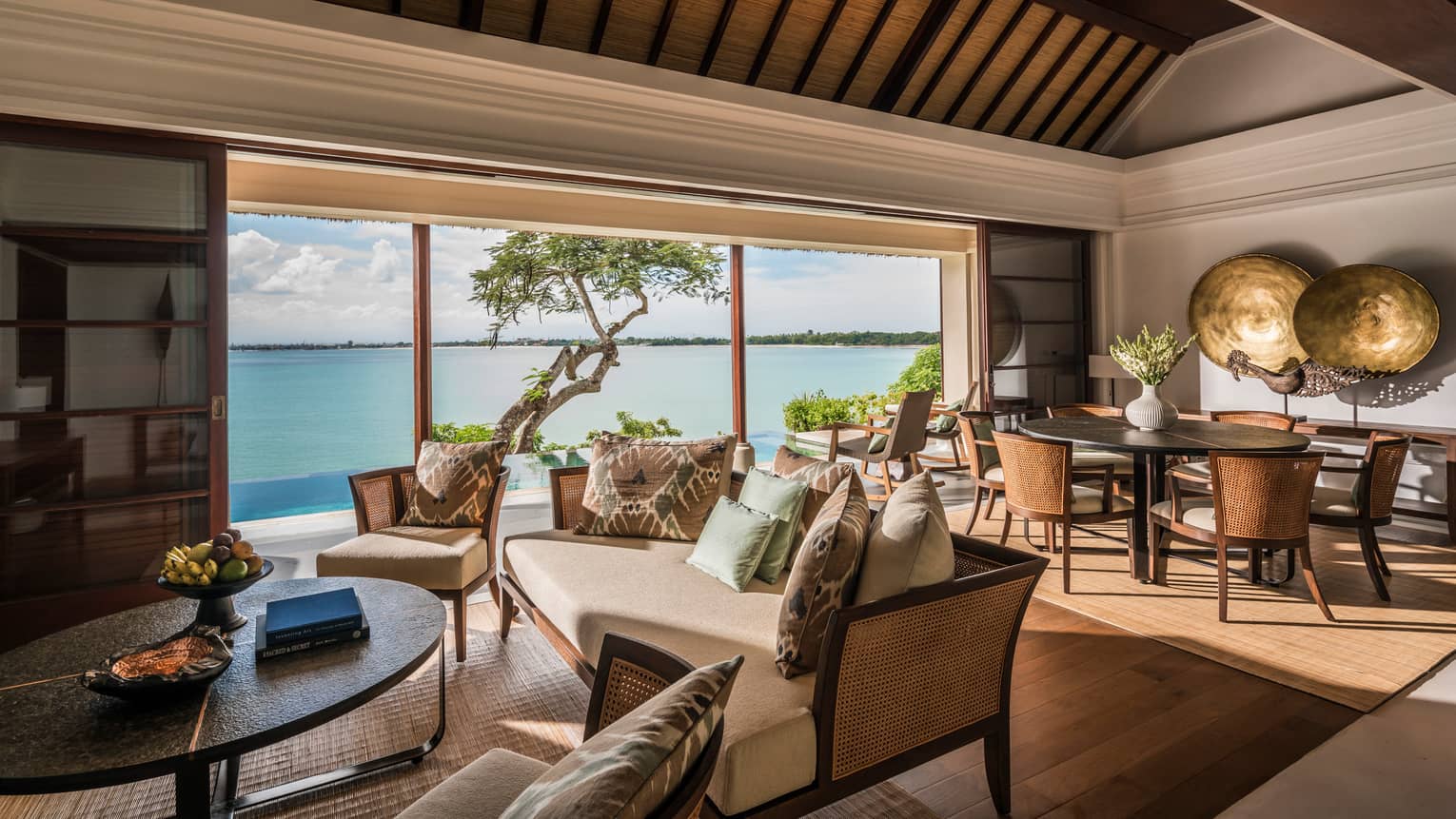 Living room with elegant wicker sofa with white cushions, dining table, large round brass plates on wall, glass wall to ocean