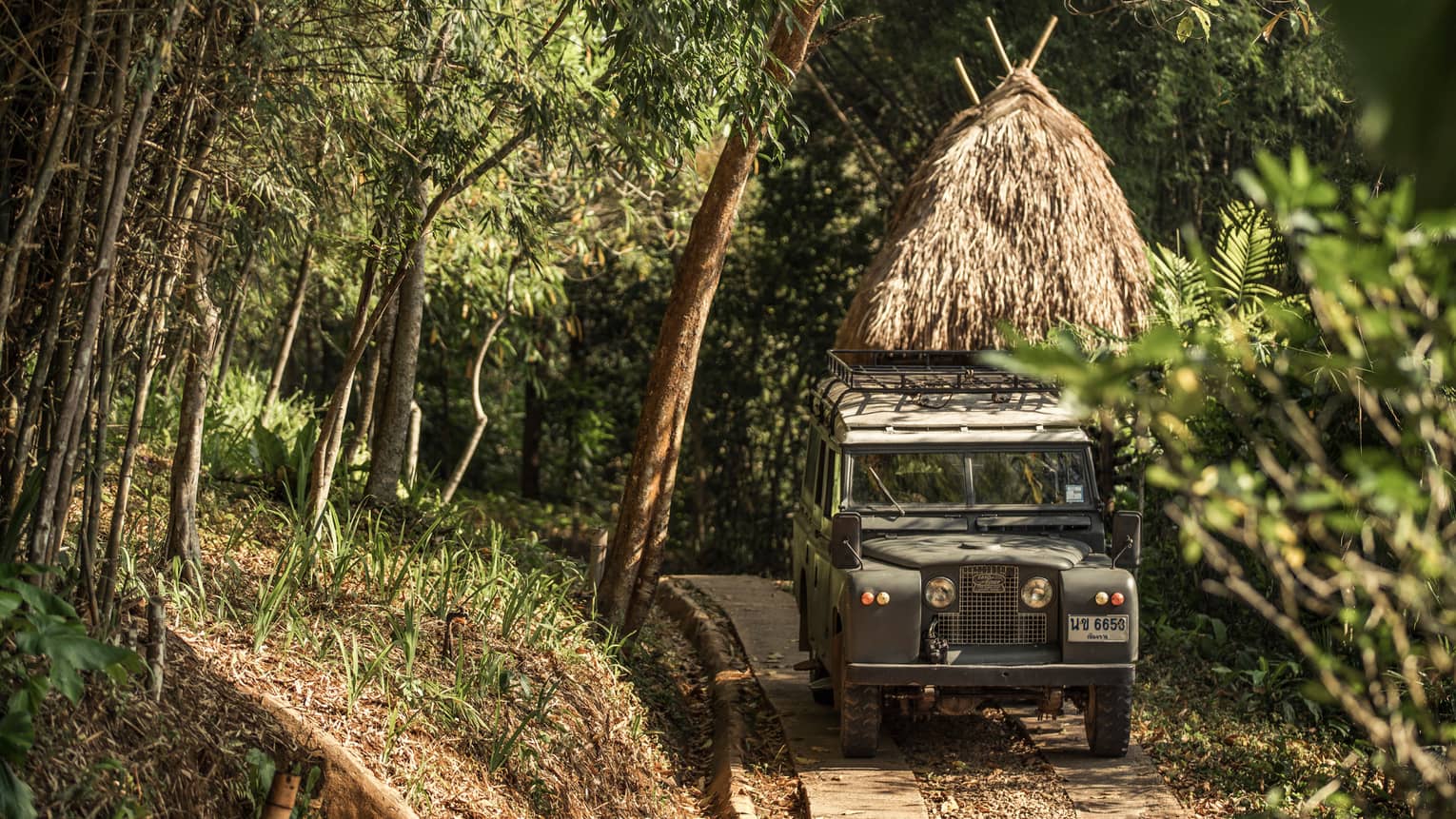 Jeep driving down path in forest past thatched-roof hut