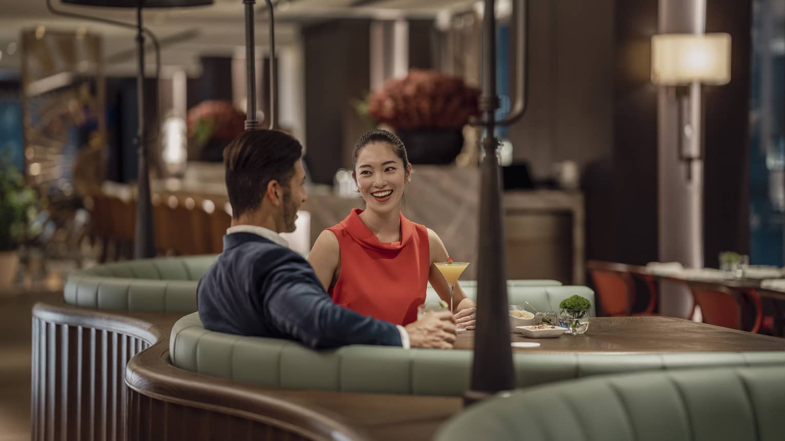 A smiling couple sit at a circular booth in an empty, dimly lit restaurant with cocktails and appetizers on their table.
