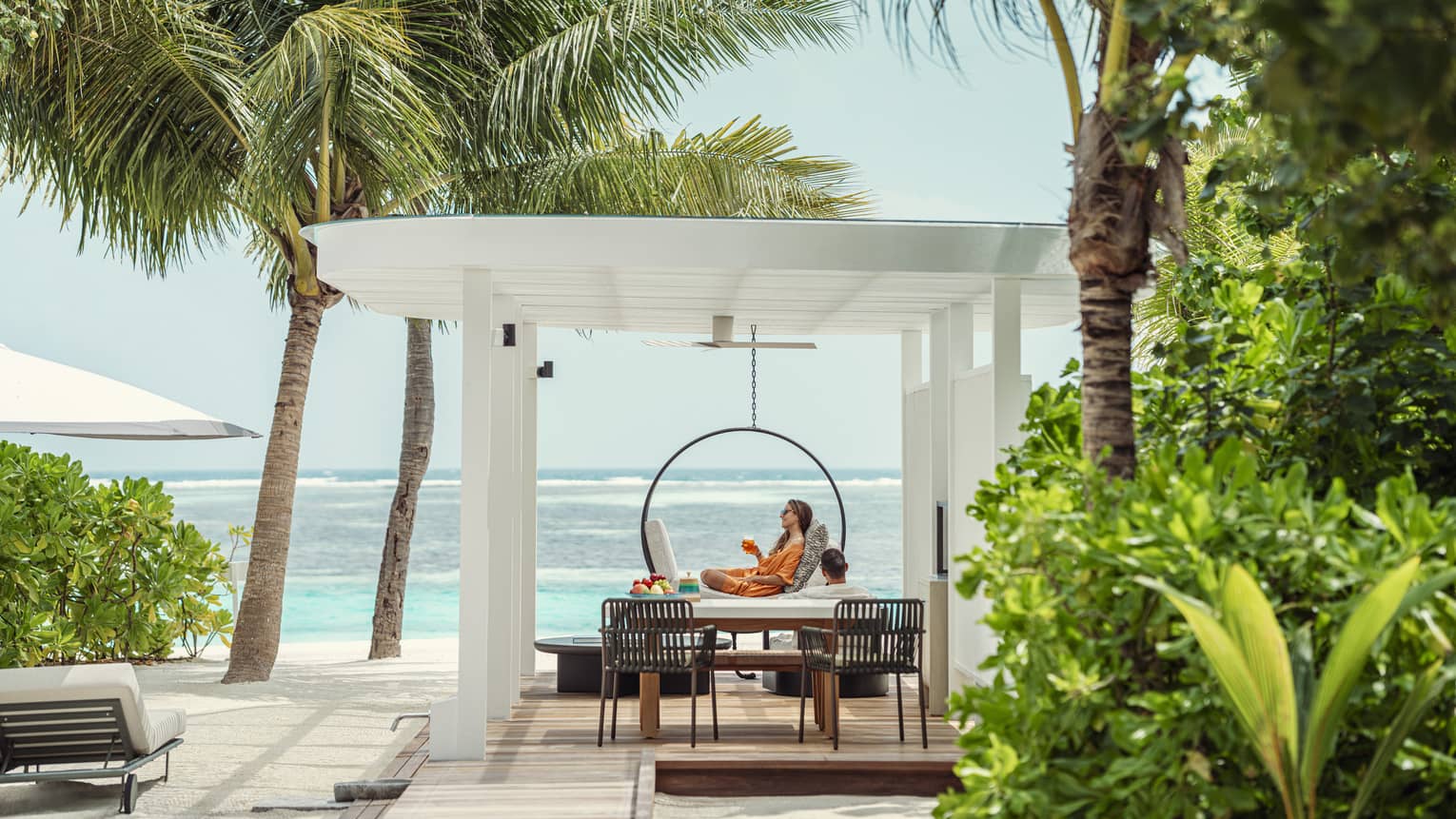 A person lounges under pergola on pool deck of private villa next to beach