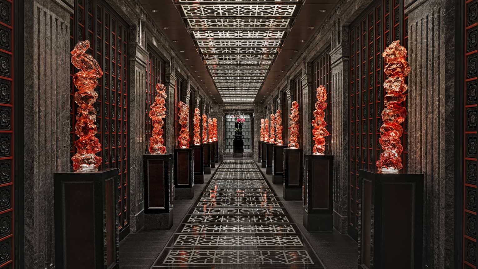 View down a long black-tiled hall lined with orange glass sculptures on pedestals, the geometric ceiling mirroring the floor.